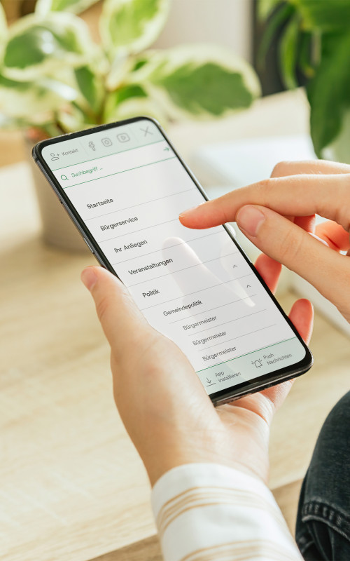 In der Hand wird ein Smarthpone gehalten, welches einen Ausschnitt des neuen Gemeindeportal Designs zeigt.