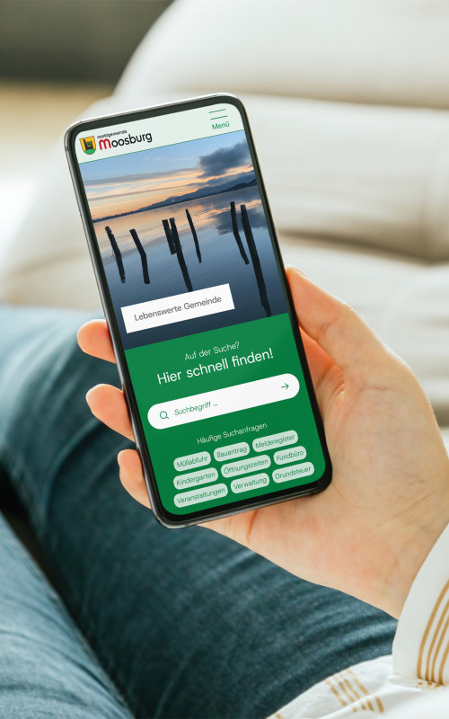 In der Hand wird ein Smarthpone gehalten, welches einen Ausschnitt des neuen Gemeindeportal Designs zeigt.